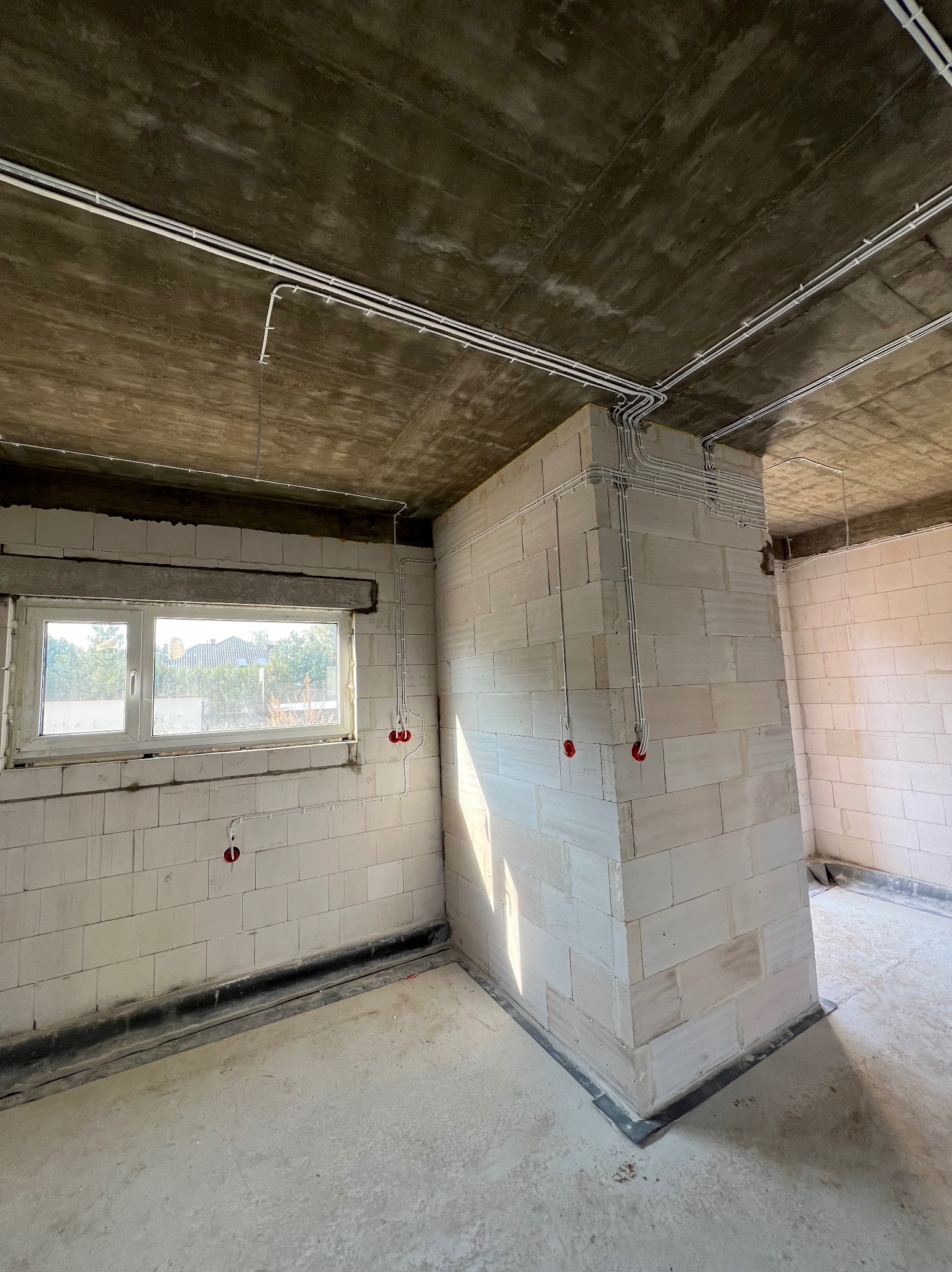 Wnętrze surowego budynku z instalacją elektryczną w białych rurkach PCV, widoczne puszki podtynkowe z czerwonymi zaślepkami, ściany z białych bloczków, surowy betonowy strop.