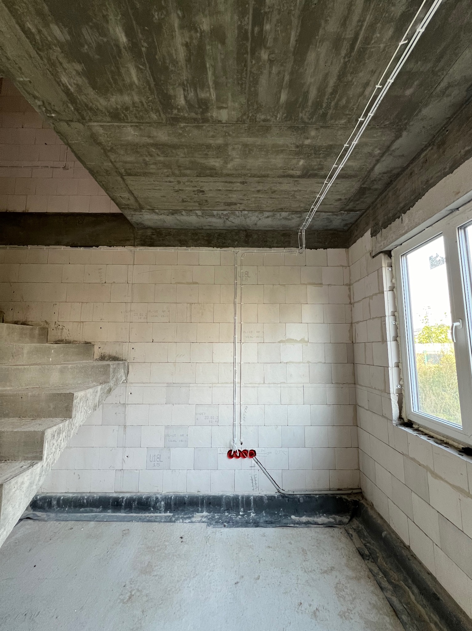 Surowy stan wnętrza z betonowymi schodami i oknem, widoczne przewody elektryczne zamocowane do ściany, czerwona puszka instalacyjna na ścianie z białych bloczków.