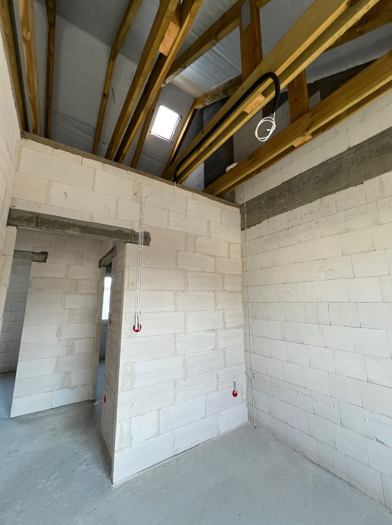 Surowy stan wnętrza z bloków, z widocznymi przewodami elektrycznymi prowadzonymi po ścianach i pod sufitem, drewniana konstrukcja dachu z oknem dachowym.