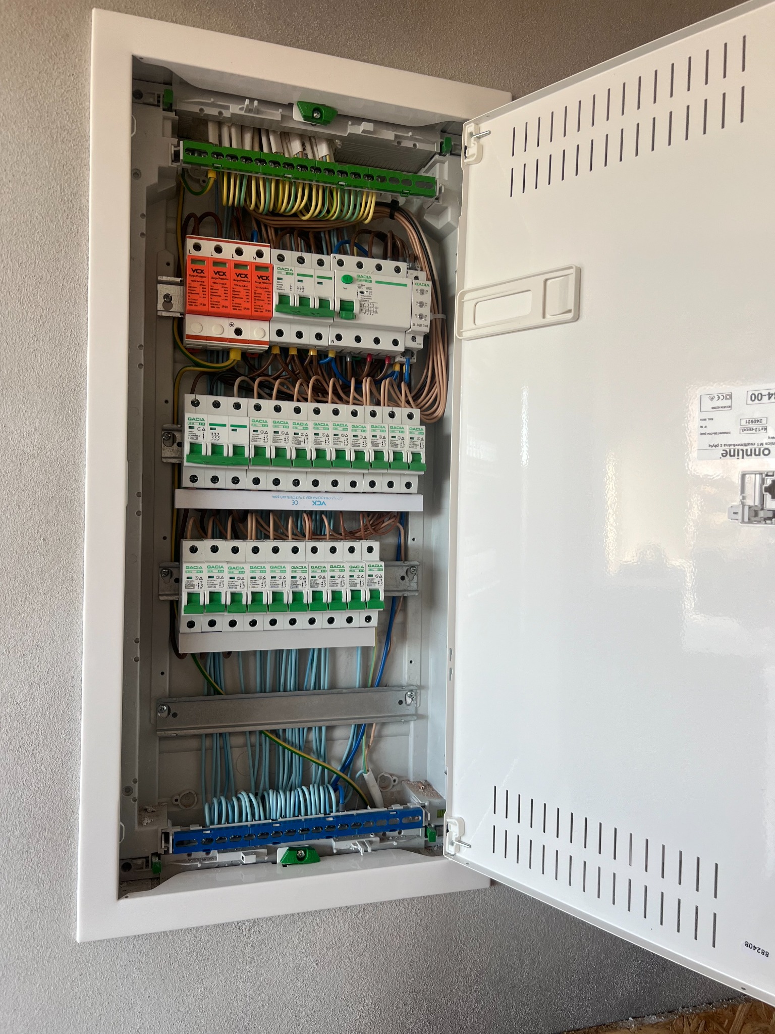 Otwarta skrzynka bezpiecznikowa z widocznymi przewodami, bezpiecznikami i ogranicznikami przepięć w kolorach zielonym, pomarańczowym i niebieskim, zamontowana na szarej ścianie.