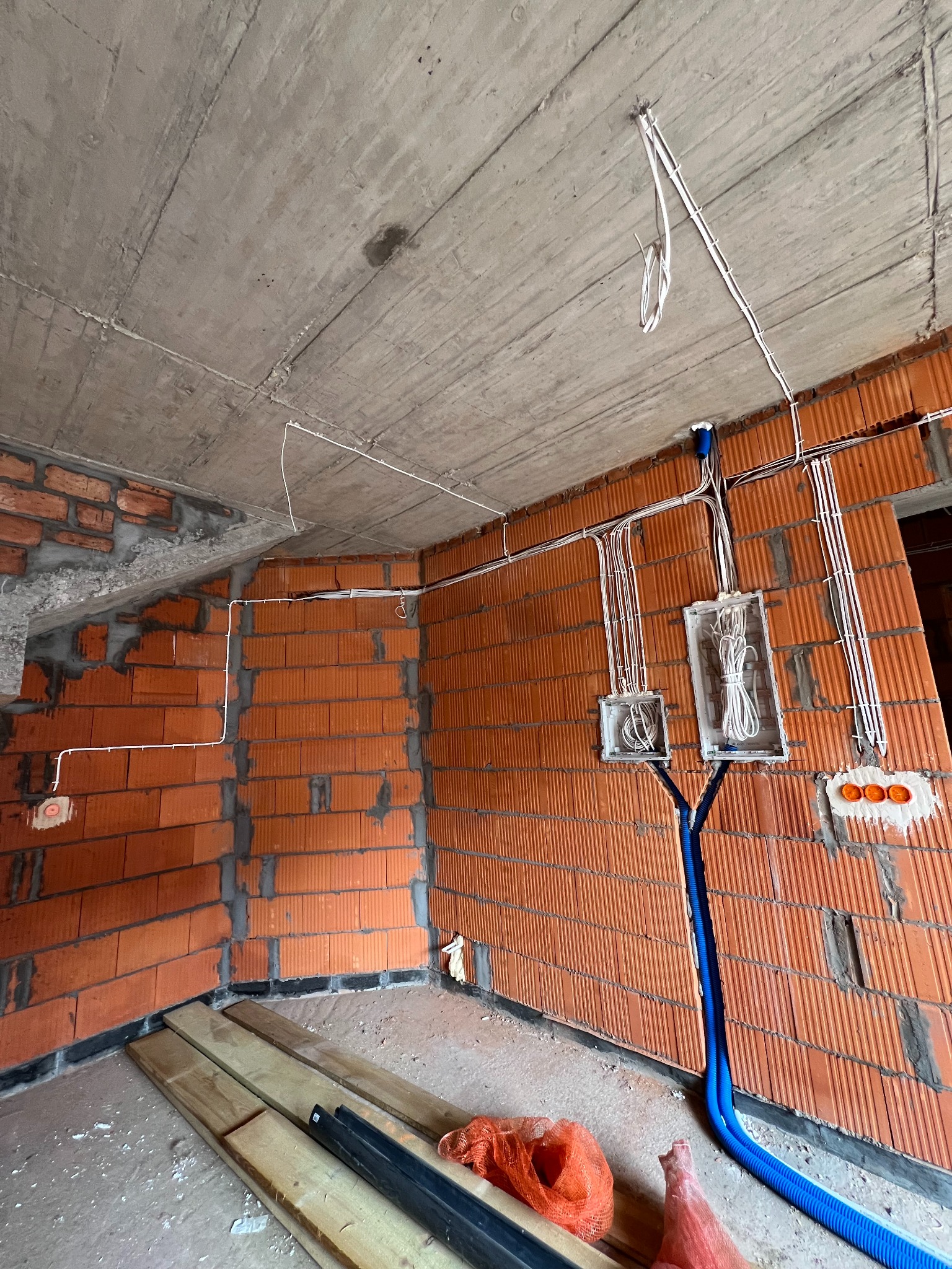 Surowy stan wnętrza z ceglanymi ścianami, betonowym stropem i rozprowadzoną instalacją elektryczną w peszlach i puszkach, widoczne kable i rury instalacyjne.