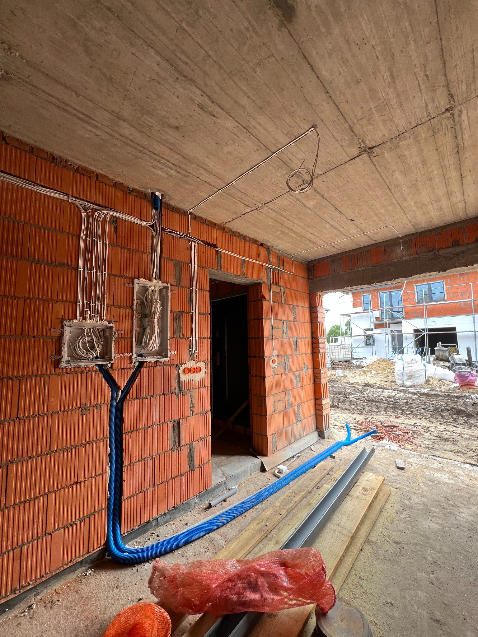 Surowy stan: instalacja elektryczna w budynku z czerwonej cegły, widoczne puszki instalacyjne, przewody w peszlach, betonowy sufit i plac budowy w tle.