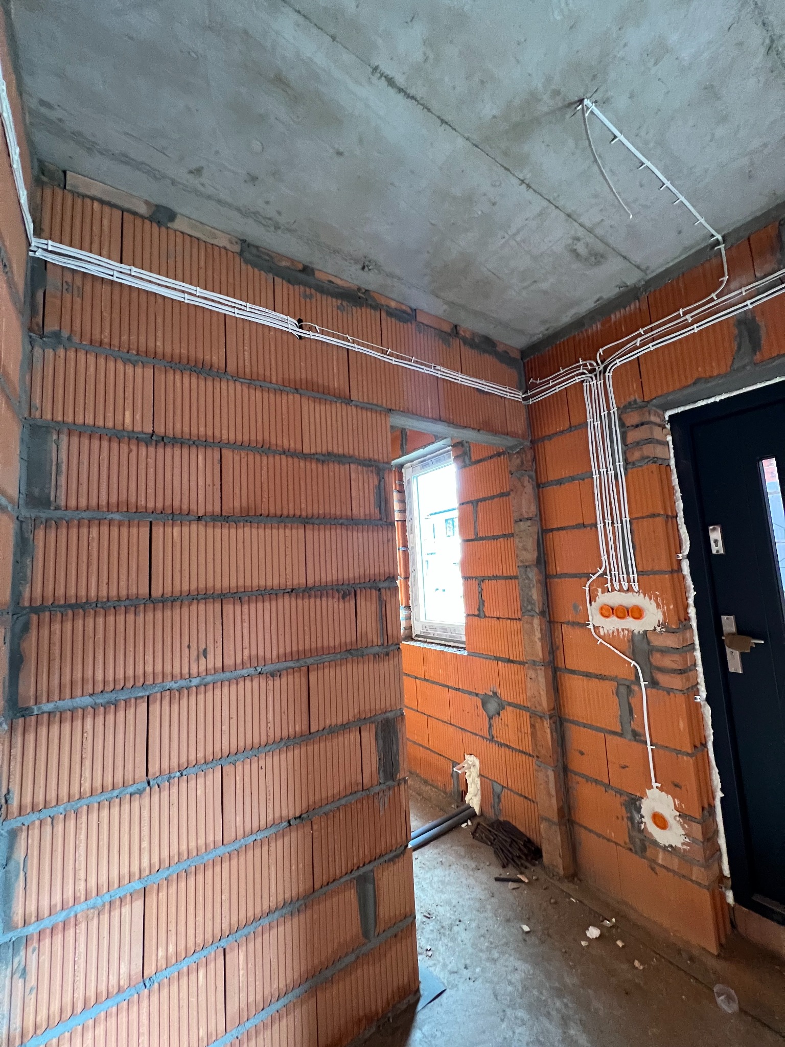 Wewnętrzne ściany z czerwonej cegły dziurawki w trakcie prac, z widocznymi białymi kablami elektrycznymi ułożonymi wzdłuż ścian i pod sufitem, oraz puszkami instalacyjnymi zalewanymi gipsem.