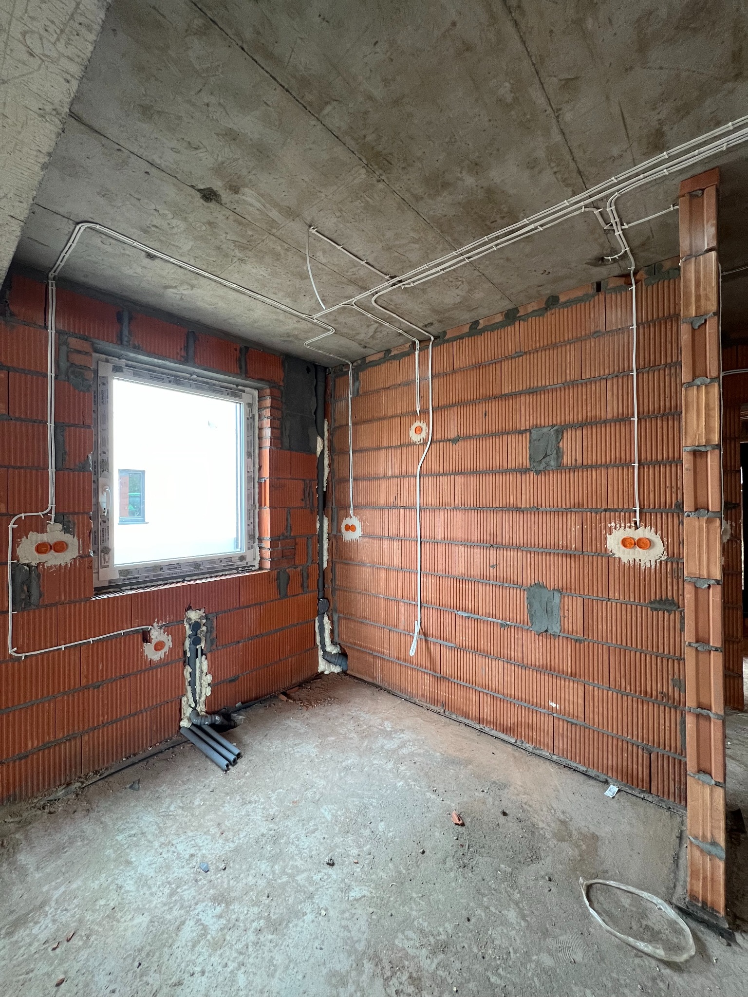 Surowy stan: wnętrze budynku w trakcie prac, widoczne kable elektryczne prowadzone po suficie i ścianach z cegły, puszki instalacyjne w ścianach, okno z widokiem na inny budynek, betonowa podłoga...