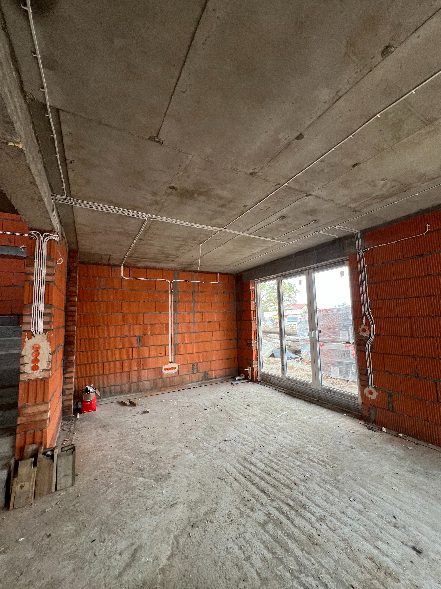 Surowy stan wnętrza z ceglanymi ścianami, widoczne białe kable elektryczne poprowadzone po suficie i ścianach, puszki elektryczne w murze, okno z widokiem na zewnątrz.