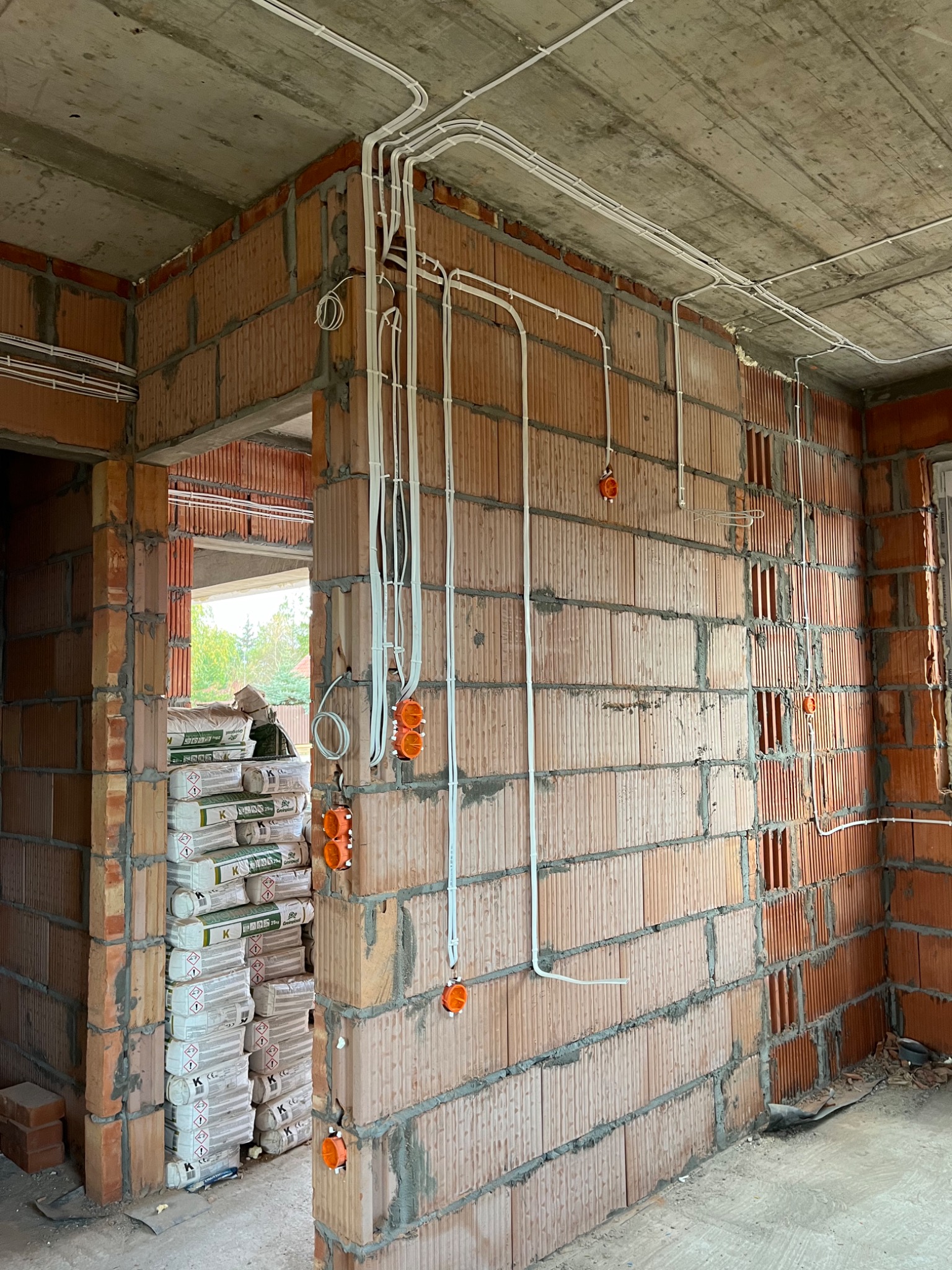 Surowy mur z cegły z widocznymi białymi rurkami instalacji elektrycznej i pomarańczowymi puszkami pod gniazdka, w tle stos worków z zaprawą budowlaną.