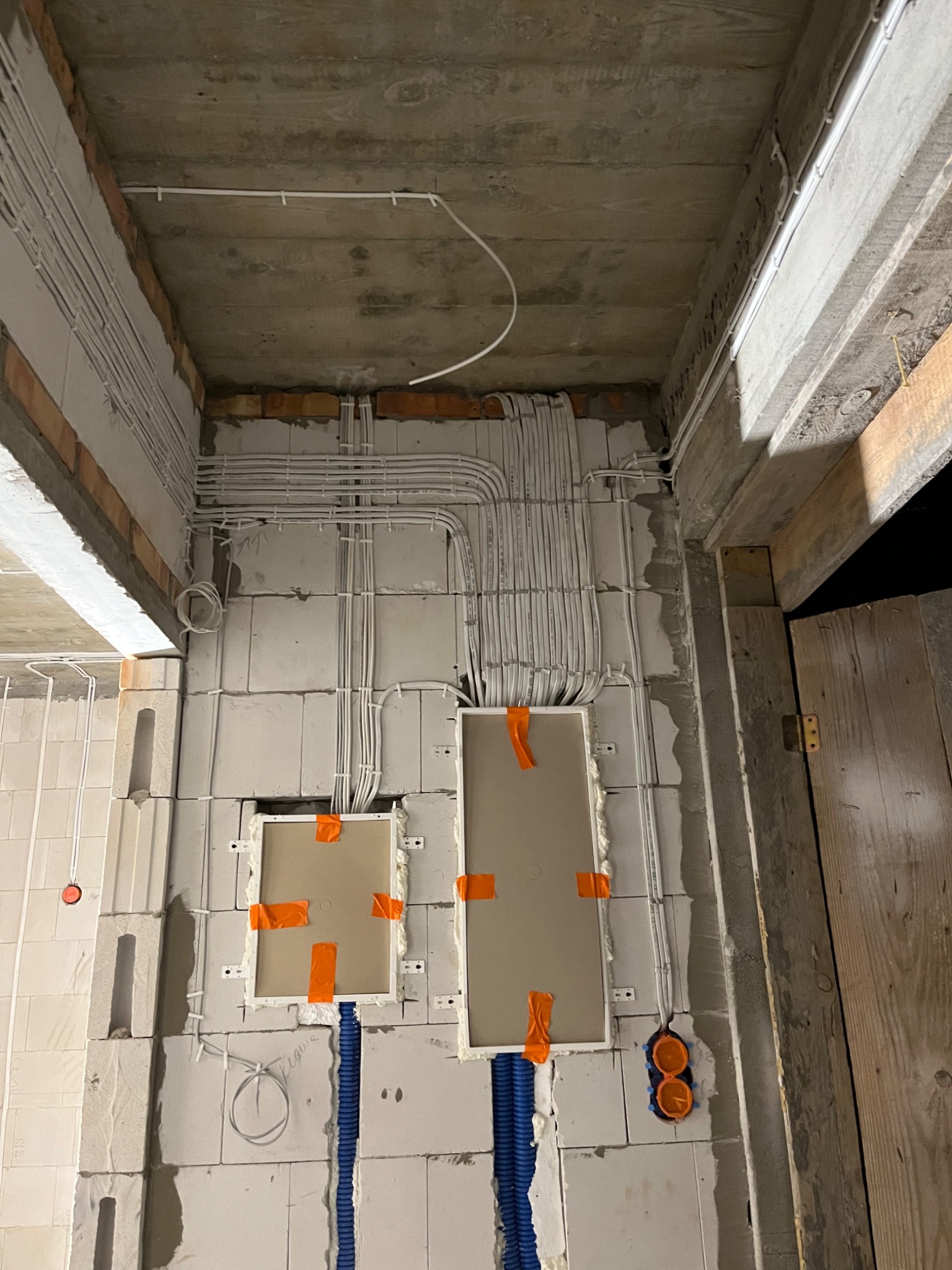 Wewnętrzne okablowanie w surowym stanie: białe przewody ułożone w korytkach na ścianie z bloków, dwa otwory na puszki elektryczne zabezpieczone pomarańczową taśmą, niebieskie rury karbowane...