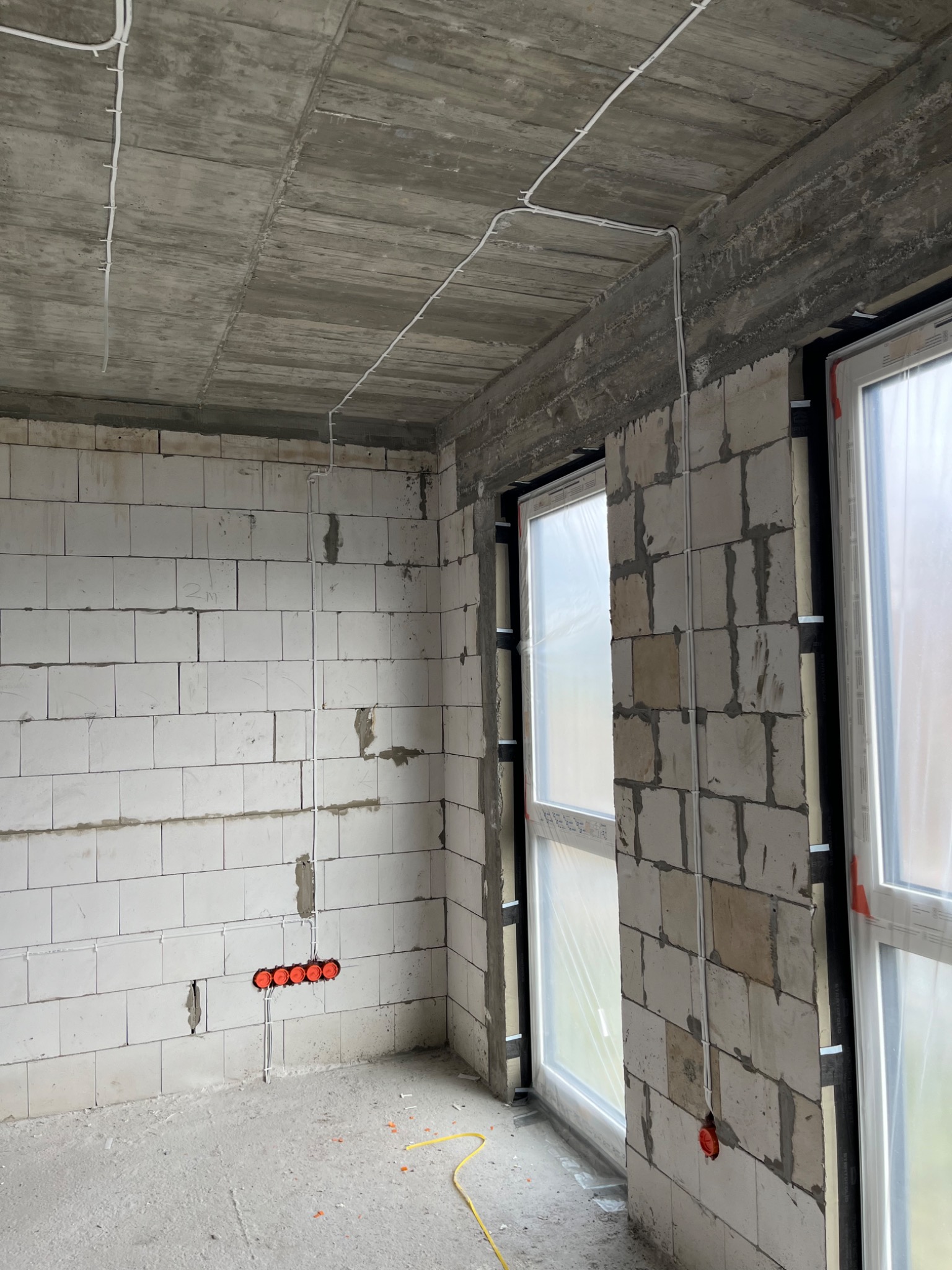 Surowy stan wnętrza z widocznymi przewodami elektrycznymi prowadzonymi po ścianach i suficie, pomarańczowe puszki instalacyjne w ścianie z betonu komórkowego, okna zabezpieczone folią.