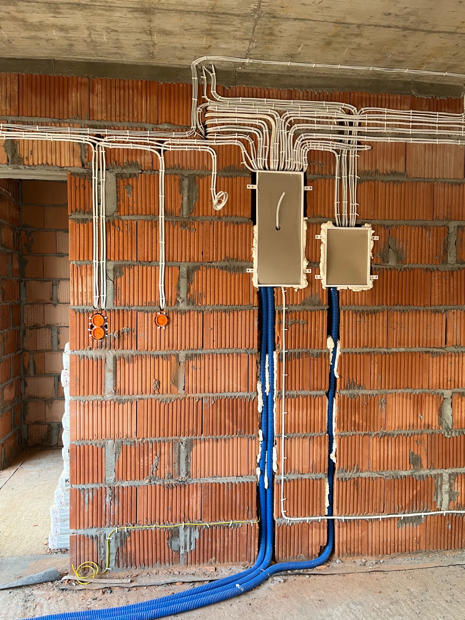 Surowa ściana z cegły z zamontowanymi puszkami elektrycznymi, białymi przewodami w peszlach ułożonymi na suficie i niebieskimi peszlami w bruzdach, widoczne pomarańczowe gniazda podtynkowe.