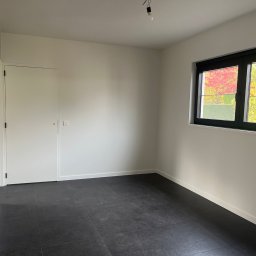 DM-domy marzeń - Pusty pokój z czarną podłogą, białymi ścianami, białymi drzwiami i oknem z czarną ramą, przez które widać jesienne drzewa.