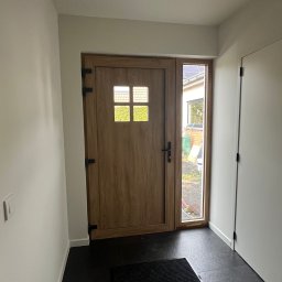 DM-domy marzeń - Nowoczesne wejście do domu w Krosno Odrzańskie z drewnianymi drzwiami z szybkami i widokiem na ogród przez boczne okno, czarna wycieraczka na ciemnej podłodze.