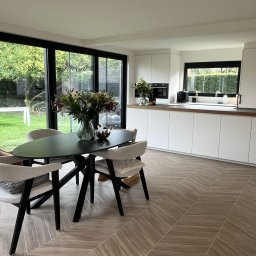 DM-domy marzeń - Nowoczesna kuchnia z jadalnią, białe szafki kuchenne, drewniany blat, okno z widokiem na ogród, stół z bukietem kwiatów, parkiet ułożony w jodełkę.