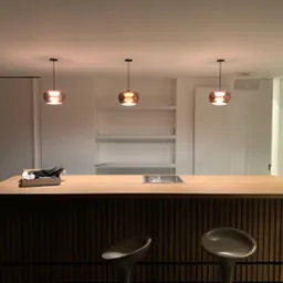 Nowoczesna wyspa kuchenna z drewnianym blatem, metalowym zlewem i ciemnym, prążkowanym frontem, oświetlona trzema miedzianymi lampami wiszącymi, z dwoma hokerami i białymi szafkami w tle.