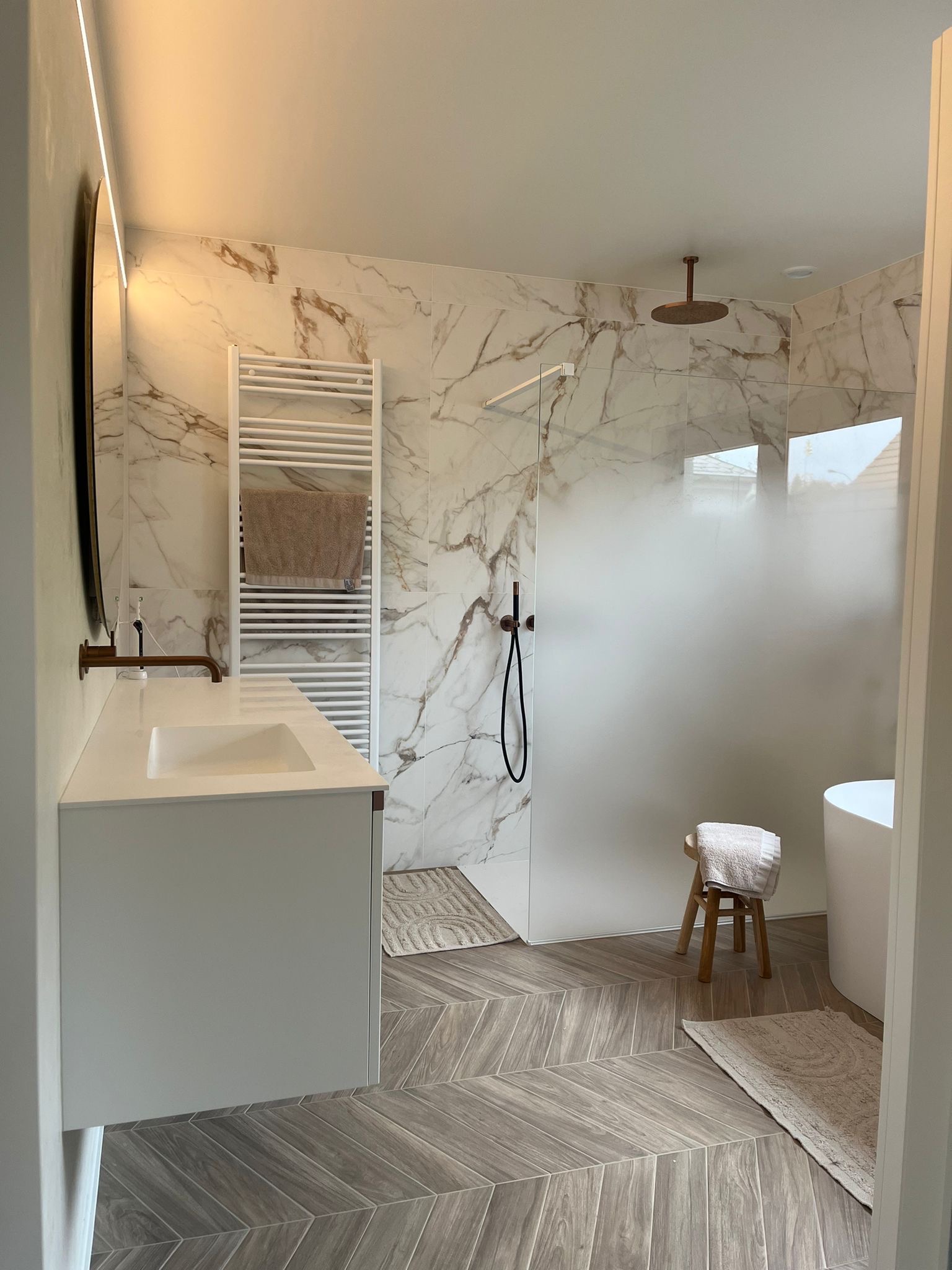 Nowoczesna łazienka z marmurowymi płytkami, kabiną prysznicową ze szklaną ścianką, umywalką na szafce, grzejnikiem drabinkowym i drewnopodobną podłogą ułożoną w jodełkę.