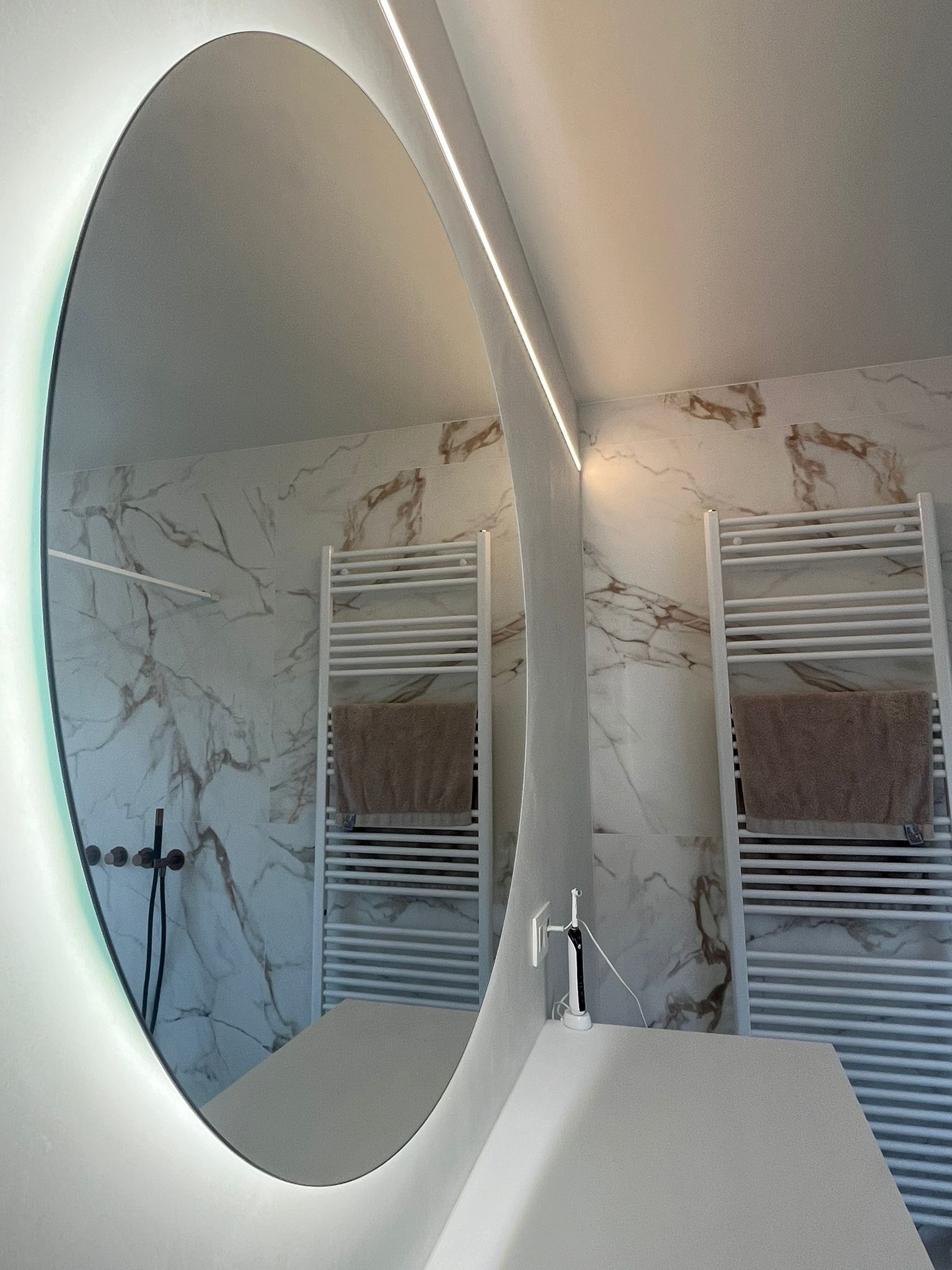 Owalne lustro z podświetleniem LED odbija marmurowe płytki, biały grzejnik drabinkowy z ręcznikiem i białą umywalkę z elektryczną szczoteczką do zębów.