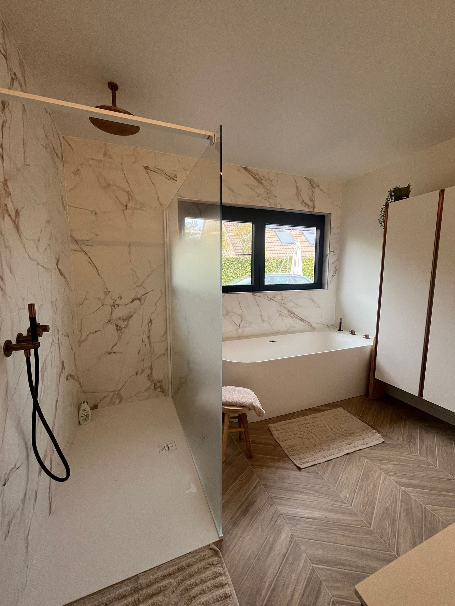 Nowoczesna łazienka z marmurowymi płytkami, kabiną prysznicową typu walk-in z deszczownicą w kolorze miedzi, białą wanną wolnostojącą i drewnianą podłogą ułożoną w jodełkę.