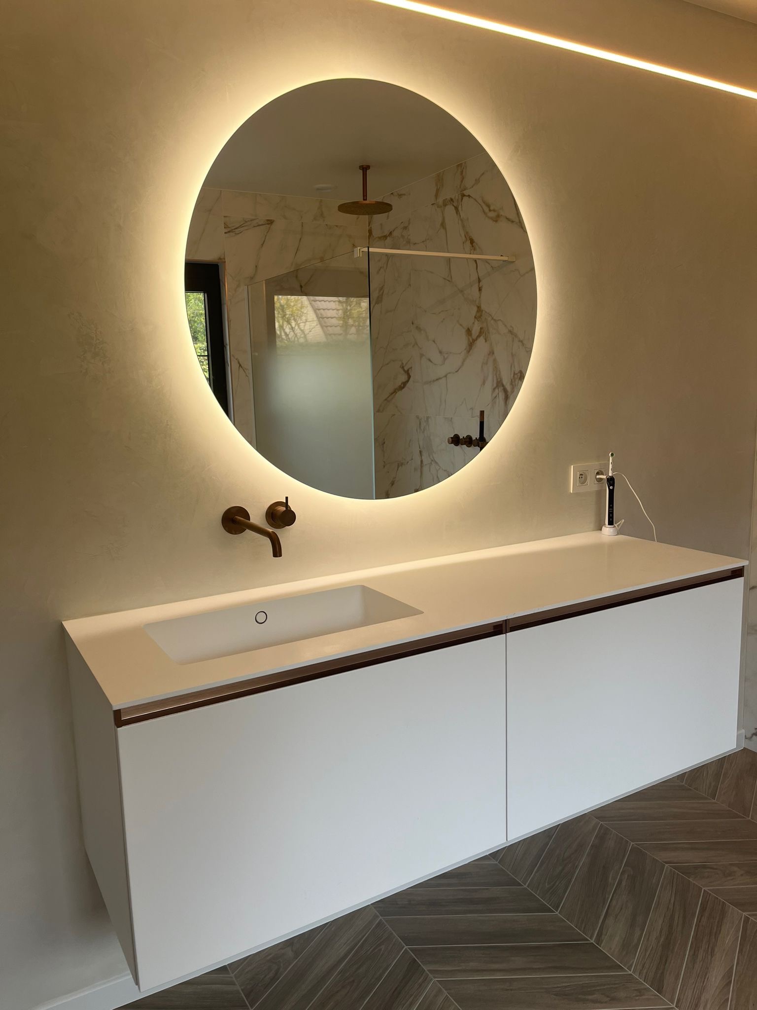 Nowoczesna łazienka z okrągłym lustrem podświetlanym LED, białą szafką pod umywalką z brązowymi akcentami i drewnopodobną podłogą ułożoną w jodełkę.