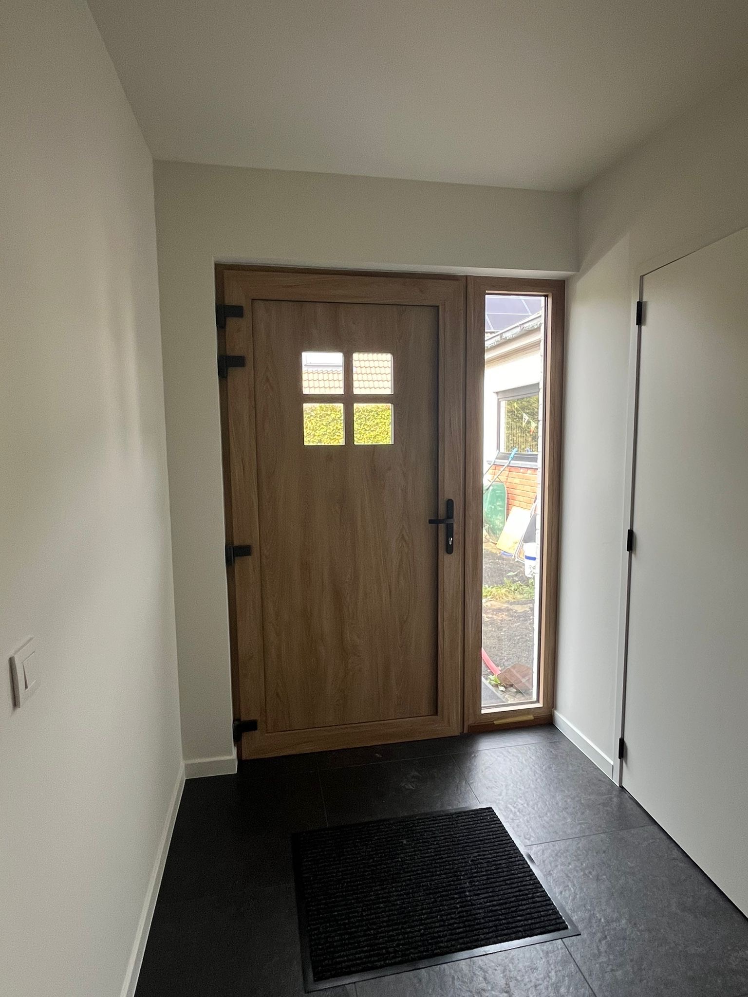 Nowoczesne wejście do domu w Krosno Odrzańskie z drewnianymi drzwiami z szybkami i widokiem na ogród przez boczne okno, czarna wycieraczka na ciemnej podłodze.