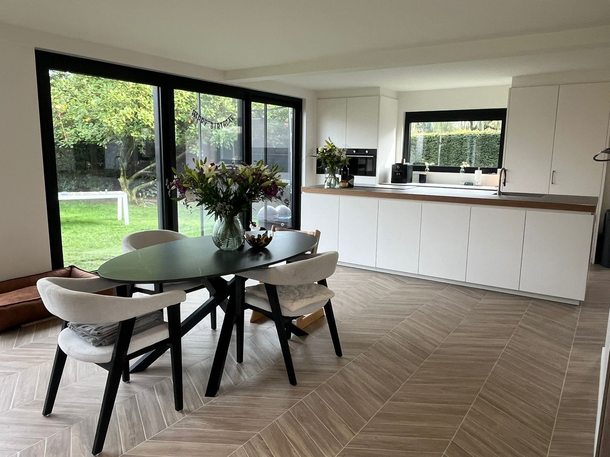 Nowoczesna kuchnia z jadalnią, białe szafki kuchenne, drewniany blat, okno z widokiem na ogród, stół z bukietem kwiatów, parkiet ułożony w jodełkę.