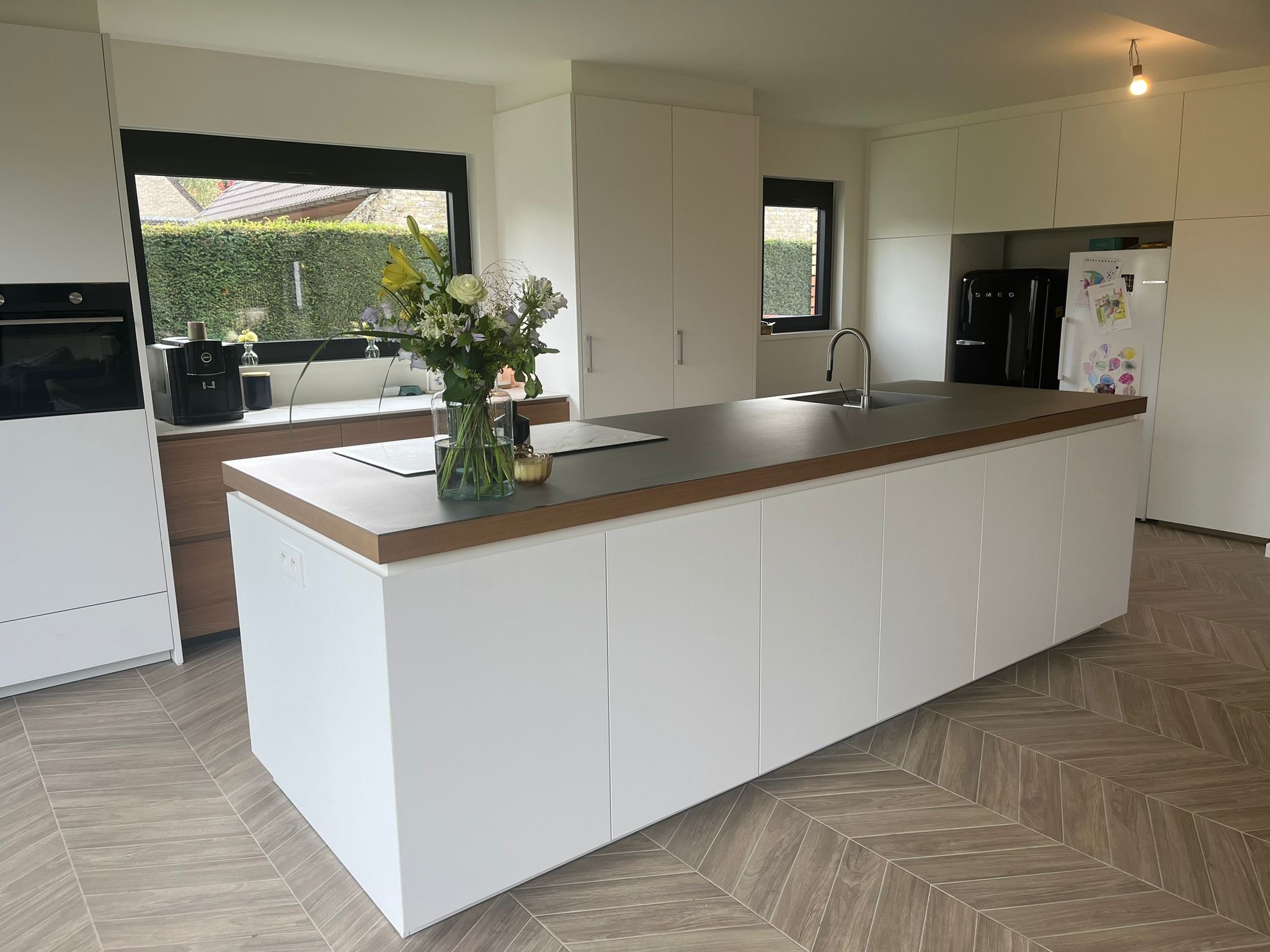 Nowoczesna kuchnia z białą wyspą, szarym blatem, drewnianym obramowaniem blatu, wzorzystą podłogą, czarną lodówką SMEG i bukietem kwiatów na wyspie.
