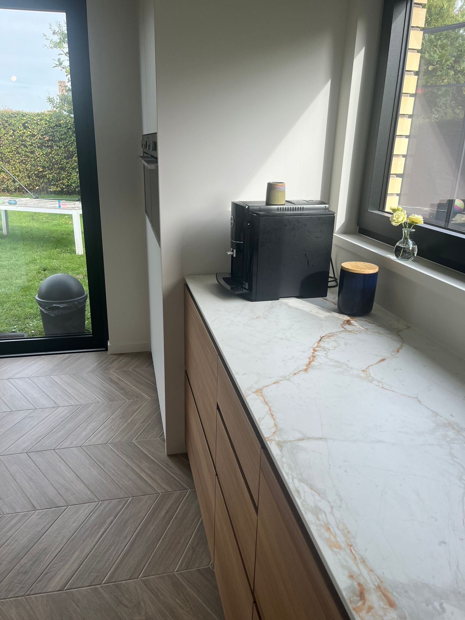 Nowoczesna kuchnia z marmurowym blatem, drewnianymi szafkami, ekspresem do kawy i podłogą w jodełkę, widok na ogród przez okno.