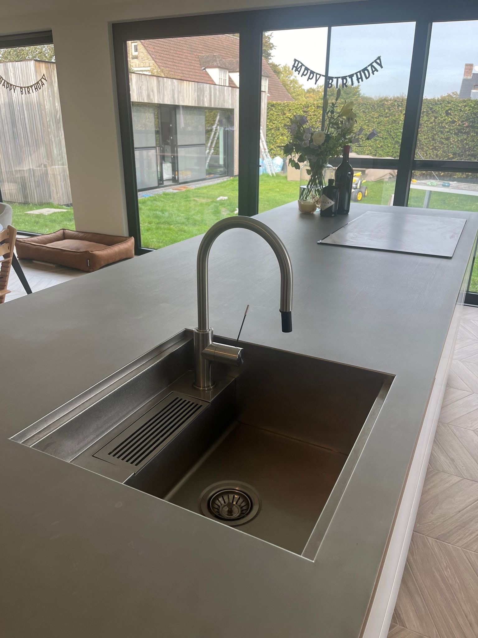 Nowoczesna, minimalistyczna kuchnia z wyspą i wbudowanym, stalowym zlewem z baterią. Za oknem widoczny ogród z drewnianym budynkiem i dekoracją urodzinową.