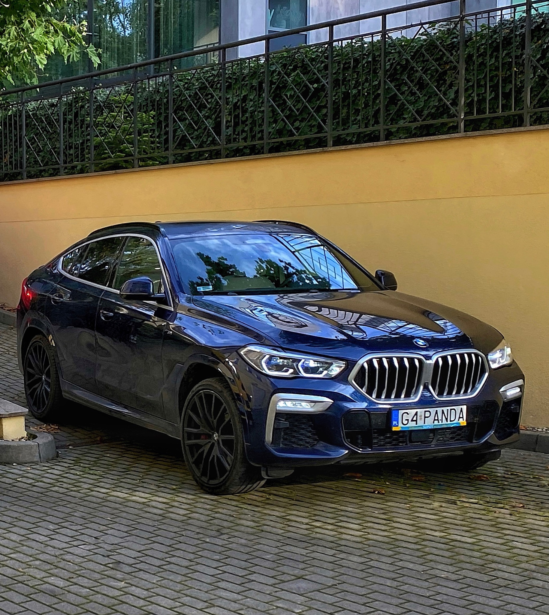 Granatowy samochód typu SUV coupe marki BMW z czarnymi felgami, zaparkowany na brukowanej nawierzchni przed żółtą ścianą z metalową kratą.