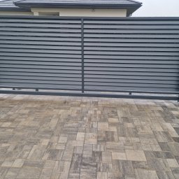 JPwelding Jakub Pawłowski - Nowoczesna, pozioma brama wjazdowa z szarych paneli, osadzona na kamiennych słupkach, z widoczną kostką brukową na podjeździe i fragmentem domu w tle.