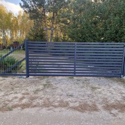 JPwelding Jakub Pawłowski - Nowoczesna, pozioma brama wjazdowa z szarego metalu, sąsiadująca z ogrodzeniem z siatki, na tle drzew i częściowo widocznego domu.