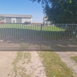 JPwelding Jakub Pawłowski - Nowoczesna, dwuskrzydłowa brama wjazdowa z pionowych, metalowych profili, prowadząca na posesję z budynkami gospodarczymi i zielonym trawnikiem w tle.