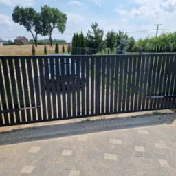 Nowoczesna, automatyczna brama wjazdowa z pionowych profili, widok z zewnątrz na posesję z polami i domami w tle.