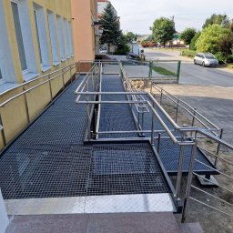 JPwelding Jakub Pawłowski - Zewnętrzna rampa z metalową kratą i nierdzewnymi poręczami, zapewniająca dostęp dla osób z niepełnosprawnościami do budynku z żółtą elewacją i białymi obramowaniami okien.