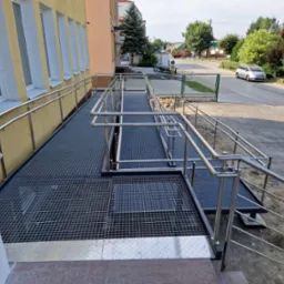 Zewnętrzna rampa z metalową kratą i nierdzewnymi poręczami, zapewniająca dostęp dla osób z niepełnosprawnościami do budynku z żółtą elewacją i białymi obramowaniami okien.