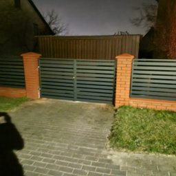 JPwelding Jakub Pawłowski - Nowoczesna brama wjazdowa z poziomych, szarych paneli, osadzona na ceglanych słupkach, oświetlona nocą, z widocznym cieniem fotografa.