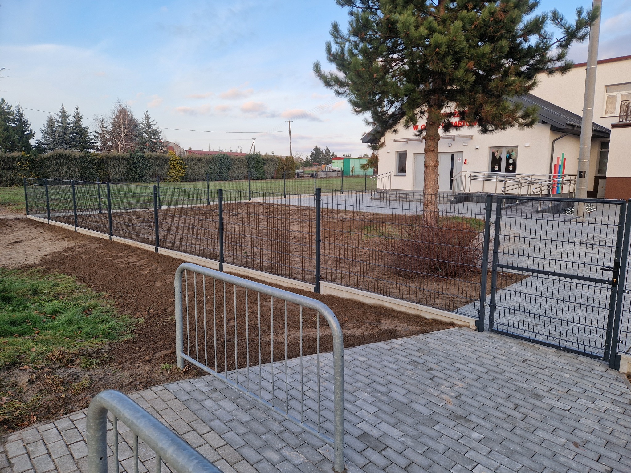Nowo postawione ogrodzenie panelowe z bramką, otaczające teren zielony przed budynkiem z drzewem iglastym, widoczne świeżo wyrównane podłoże pod trawnik.