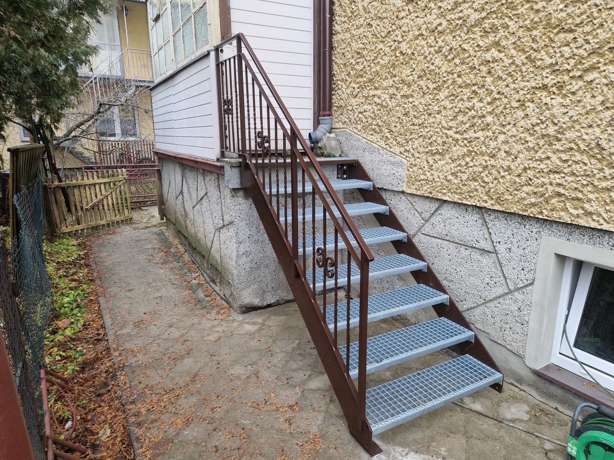 Zewnętrzne schody metalowe z brązową balustradą i kratowymi stopniami, prowadzące do wejścia budynku z elewacją z fakturą baranka, widoczne z poziomu podwórka.