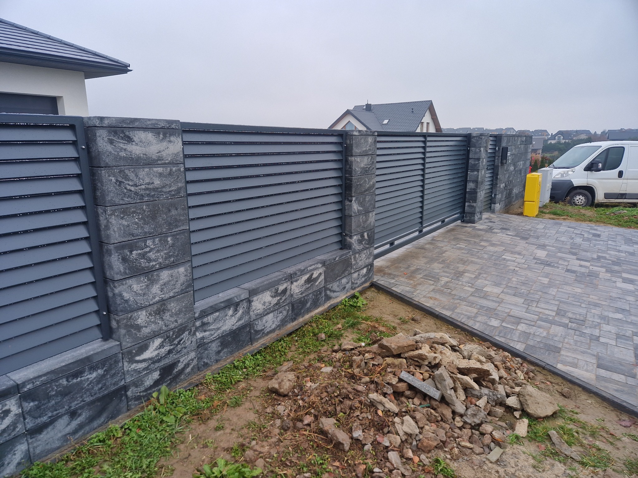 Nowoczesne ogrodzenie panelowe z bramą przesuwną, szare panele poziome, słupki z bloczków imitujących kamień, częściowo ułożona kostka brukowa, widoczny stos gruzu budowlanego.