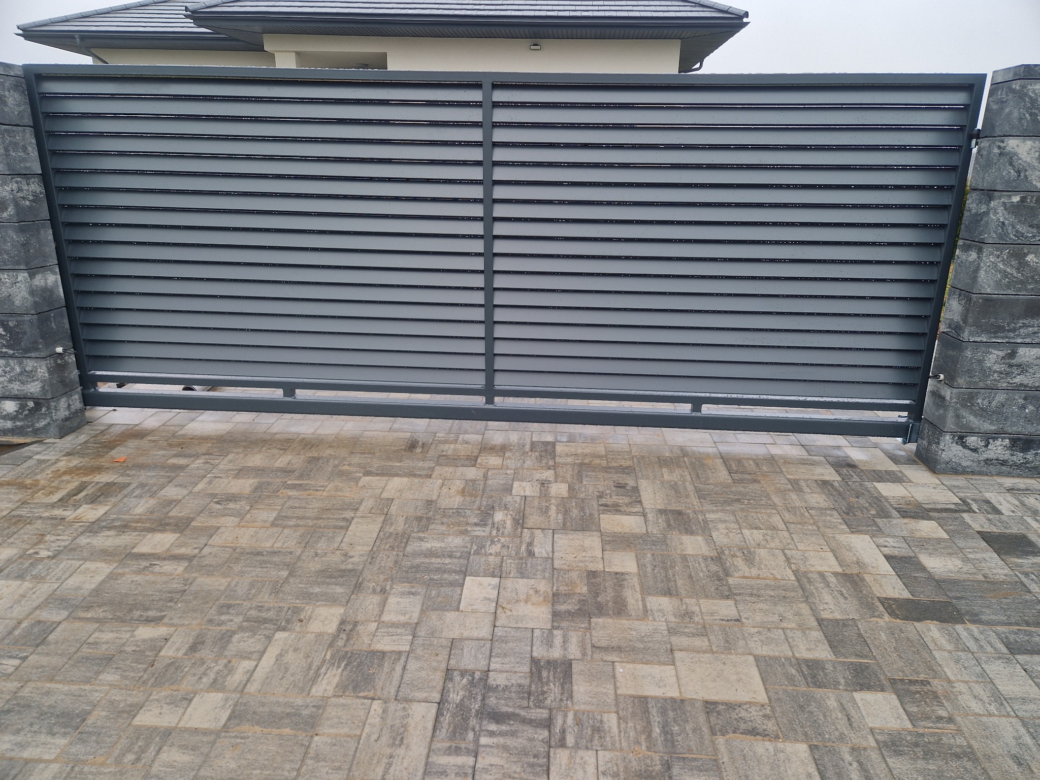 Nowoczesna, pozioma brama wjazdowa z szarych paneli, osadzona na kamiennych słupkach, z widoczną kostką brukową na podjeździe i fragmentem domu w tle.