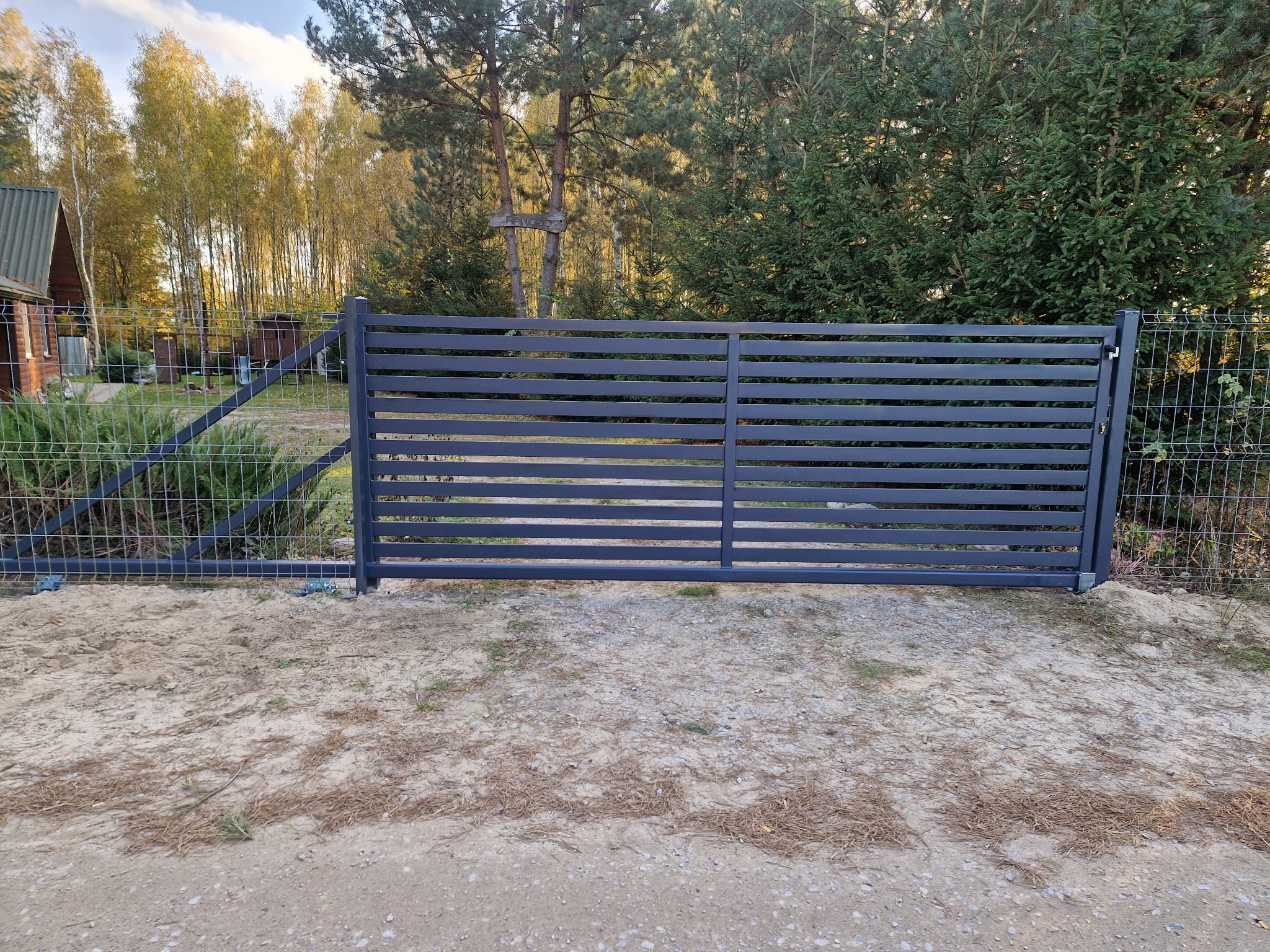 Nowoczesna, pozioma brama wjazdowa z szarego metalu, sąsiadująca z ogrodzeniem z siatki, na tle drzew i częściowo widocznego domu.