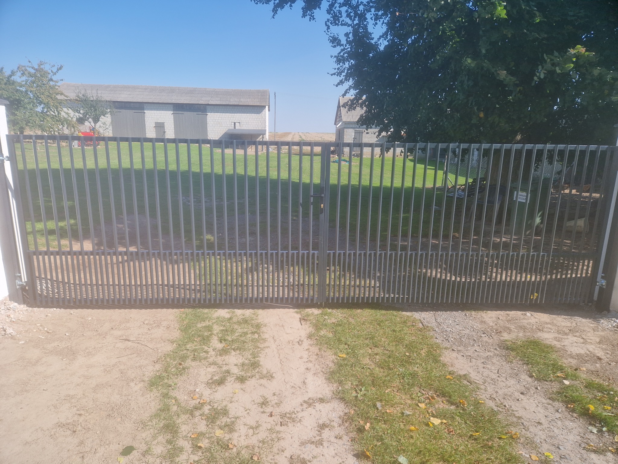 Nowoczesna, dwuskrzydłowa brama wjazdowa z pionowych, metalowych profili, prowadząca na posesję z budynkami gospodarczymi i zielonym trawnikiem w tle.