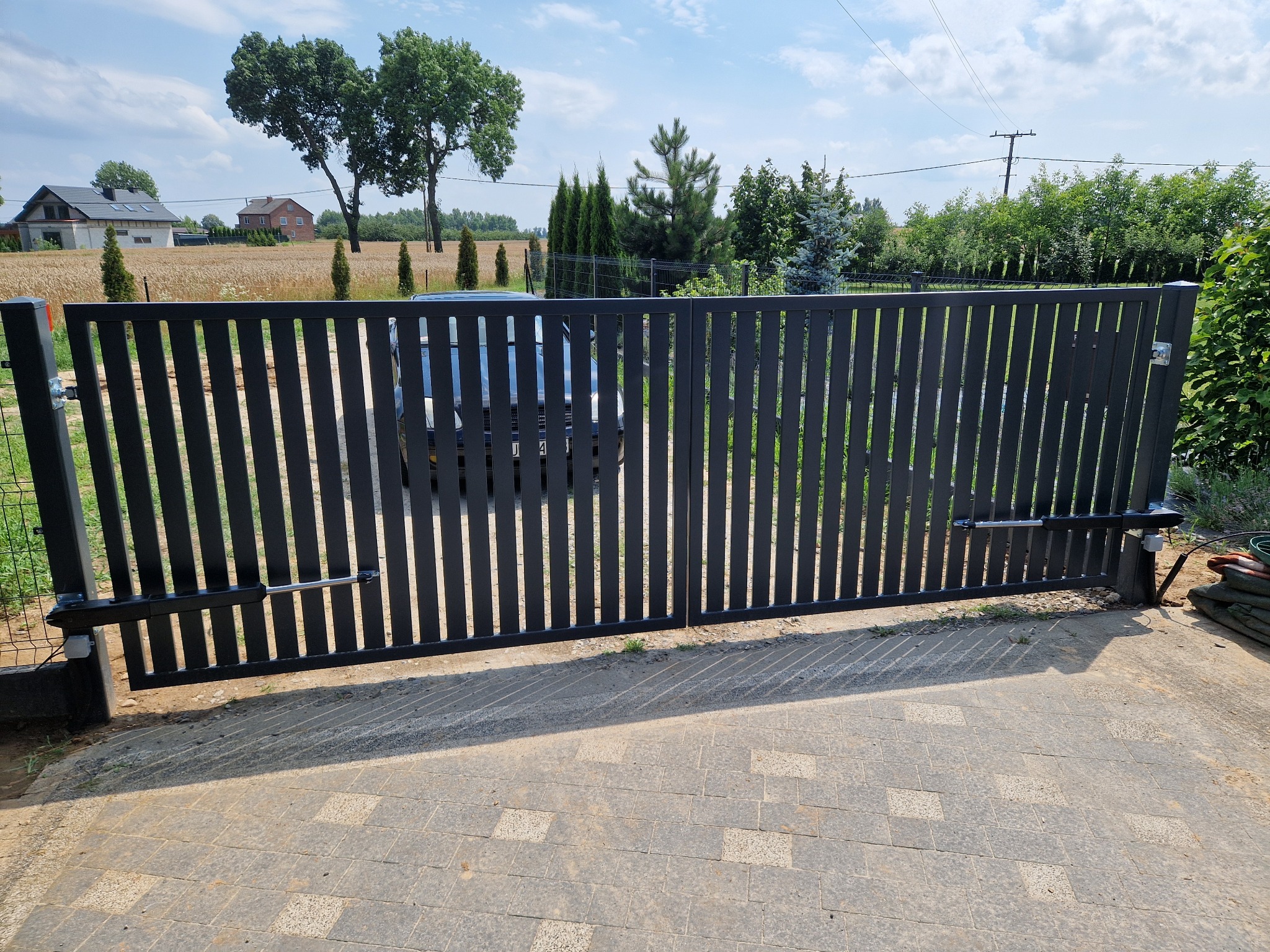 Nowoczesna, automatyczna brama wjazdowa z pionowych profili, widok z zewnątrz na posesję z polami i domami w tle.