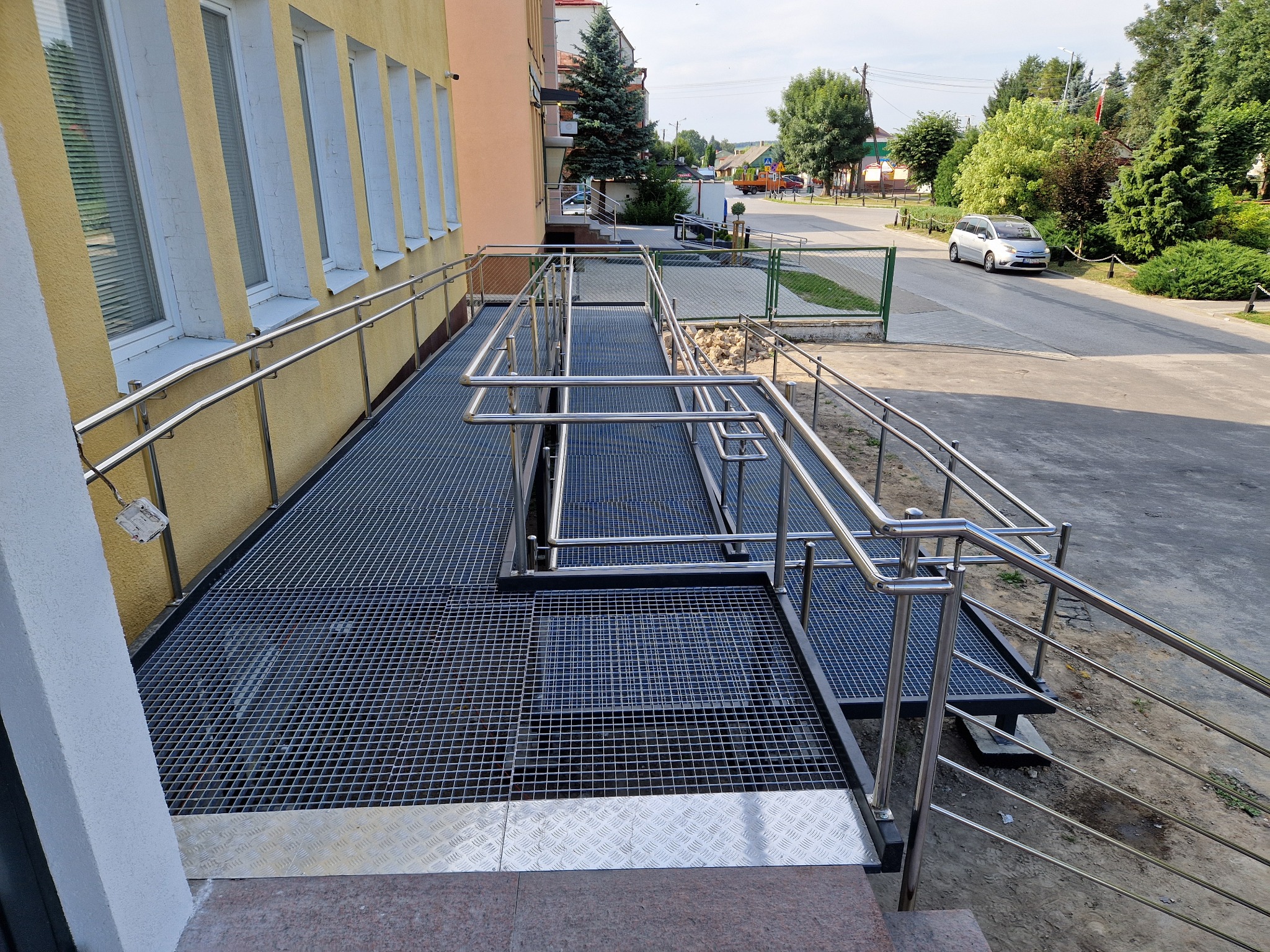 Zewnętrzna rampa z metalową kratą i nierdzewnymi poręczami, zapewniająca dostęp dla osób z niepełnosprawnościami do budynku z żółtą elewacją i białymi obramowaniami okien.