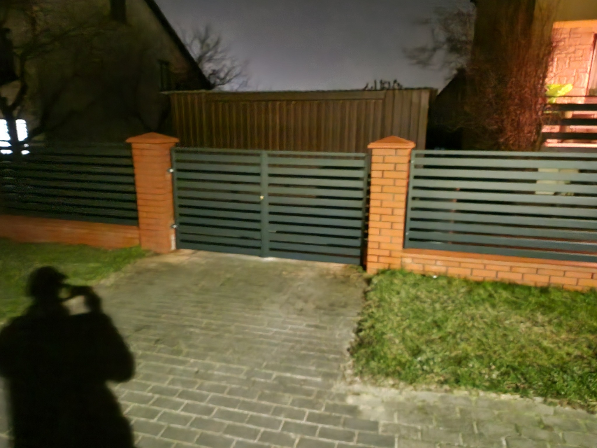 Nowoczesna brama wjazdowa z poziomych, szarych paneli, osadzona na ceglanych słupkach, oświetlona nocą, z widocznym cieniem fotografa.