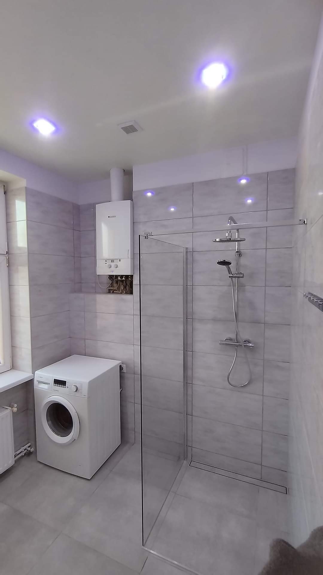Wyremontowana łazienka z kabiną prysznicową typu walk-in, pralką i piecem gazowym, płytki w odcieniach szarości, oświetlenie punktowe w suficie.