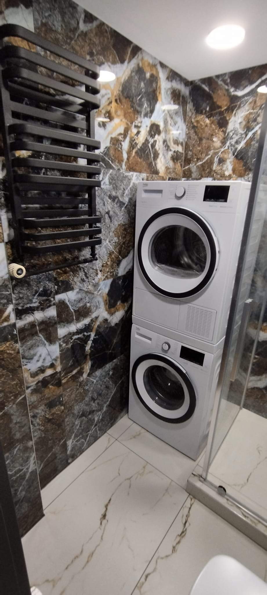 Ustawiona pionowo pralka i suszarka w łazience z marmurowymi płytkami na ścianach i podłodze, obok czarny grzejnik drabinkowy i szklana kabina prysznicowa.