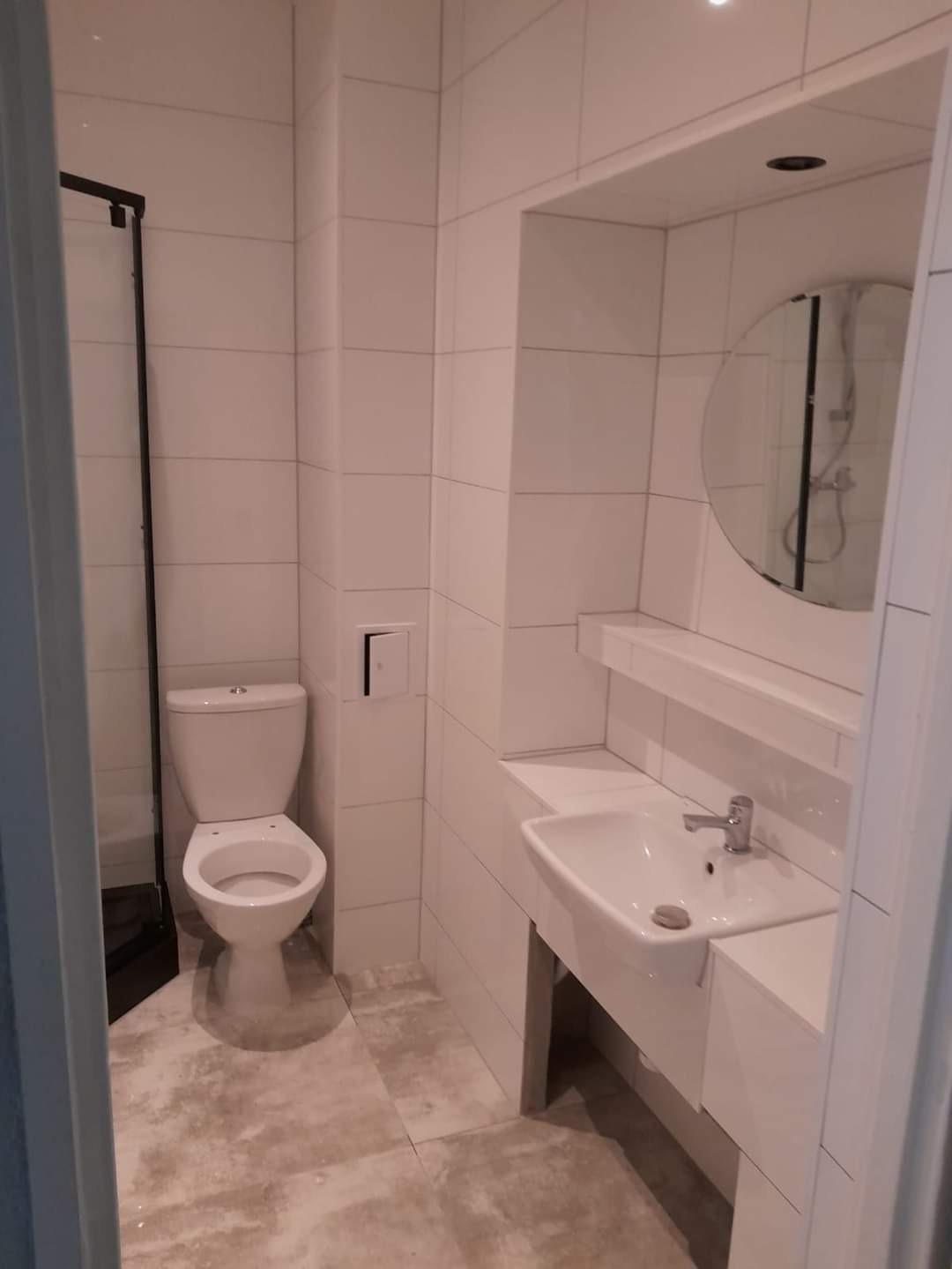 Jasna łazienka z białymi płytkami, toaleta z otwartą klapą, umywalka z okrągłym lustrem i kabina prysznicowa z czarną ramą.