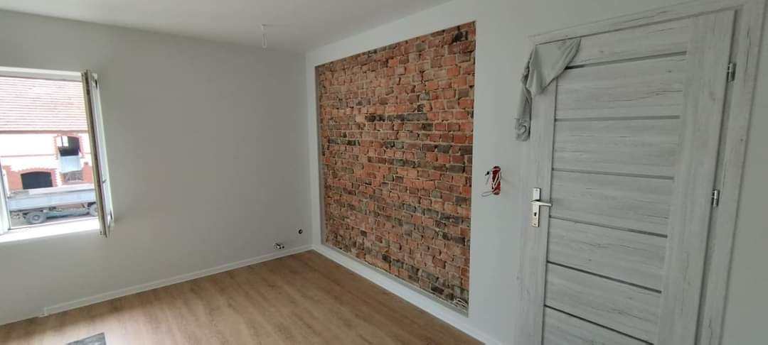 Pokój po remoncie: ściany pomalowane na biało, jedna ściana z imitacją cegły, jasna drewniana podłoga, szare drzwi z poziomymi żłobieniami, widok na budynek przez okno.
