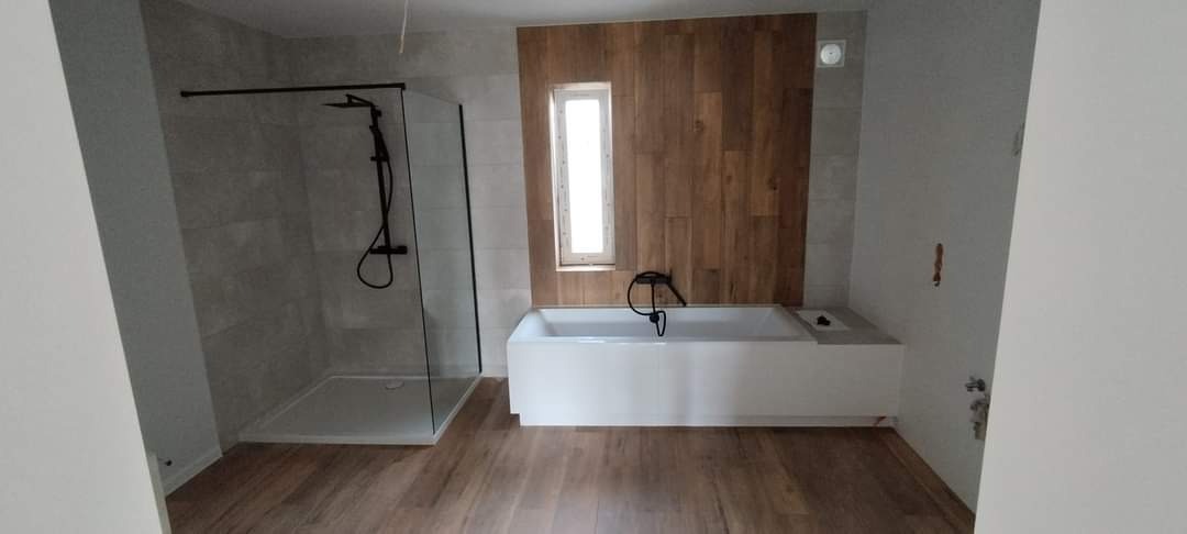 Nowoczesna łazienka z kabiną prysznicową typu walk-in z czarną armaturą, białą wanną obudowaną płytkami imitującymi drewno oraz podłogą z paneli drewnopodobnych, widok z perspektywy wejścia.