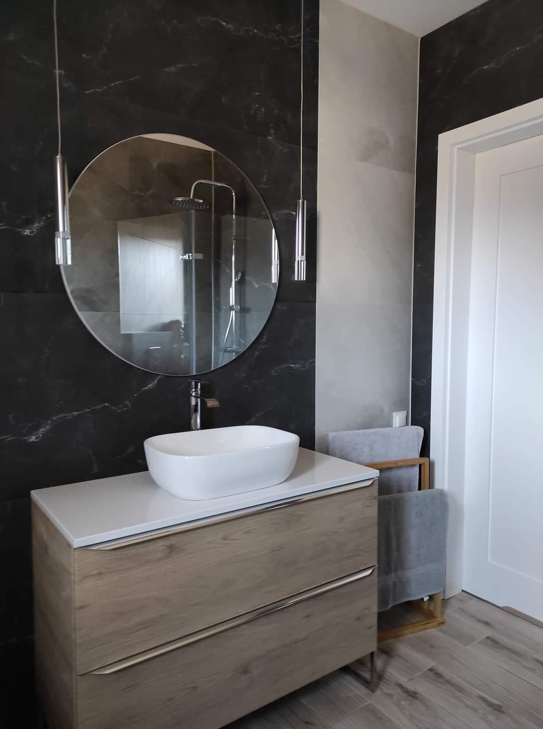 Nowoczesna łazienka z umywalką nablatową na drewnianej szafce, okrągłym lustrem i czarno-szarymi płytkami imitującymi marmur.