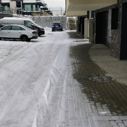 KSAL GREEN Pielęgnacja Ogrodów Kamil Kozioł - Zimowy widok na ośnieżony parking osiedlowy z widocznymi śladami opon, budynkami w tle i fragmentem podcieni. Pochmurne niebo.